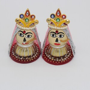 May include: Two decorative figurines of women with painted faces, wearing gold crowns and veils, and adorned with beads and rhinestones. The figurines are standing on red bases.