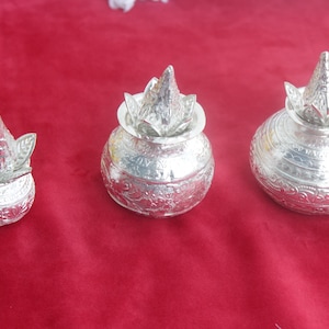 May include: Three silver-colored, ornate pots with conical lids and floral designs. The pots vary in size, with the smallest on the left and the largest on the right. They are set against a deep red background.