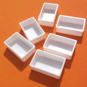 May include: Six small, rectangular, white plastic paint palettes are arranged on an orange surface. Each palette has a shallow, open compartment. The palettes are empty and ready for use.