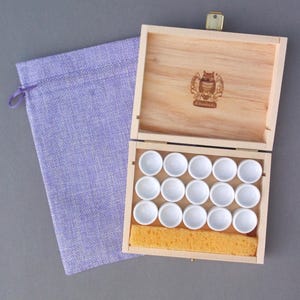 May include: A wooden paint box with a hinged lid, containing 12 white ceramic paint wells and a yellow sponge. A lavender drawstring bag is next to the box. The box lid has a logo with an owl.