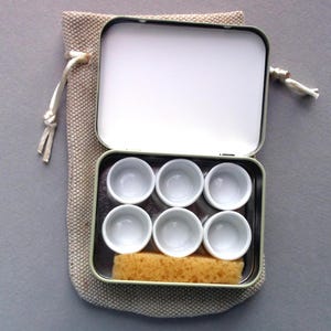 May include: A small, open metal tin containing six white ceramic paint wells and a rectangular sponge. The tin sits inside a beige drawstring bag. The lid of the tin is open, revealing a white interior.