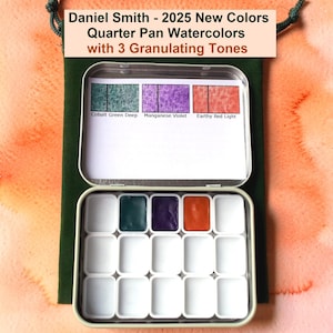 May include: An open metal watercolor paint set with three filled pans in green, purple, and orange. The set is labeled "Daniel Smith - 2025 New Colors Quarter Pan Watercolors with 3 Granulating Tones". The set is on a green pouch against an orange watercolor background.