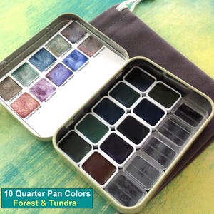 May include: Open metal watercolor paint set with 10 quarter pan colors in a variety of forest and tundra hues. The set includes a range of greens, blues, purples, and browns. A gray drawstring bag is next to the paint set. The text "10 Quarter Pan Colors Forest & Tundra" is at the bottom.