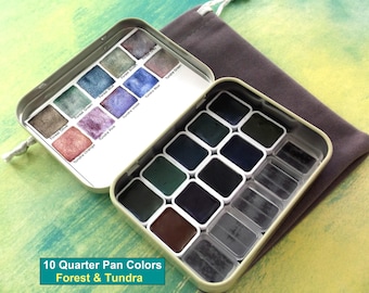 May include: Open metal watercolor paint set with 10 quarter pan colors in a variety of forest and tundra hues. The set includes a range of greens, blues, purples, and browns. A gray drawstring bag is next to the paint set. The text "10 Quarter Pan Colors Forest & Tundra" is at the bottom.