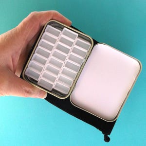 May include: Open metal watercolor paint box with multiple empty white paint pans. The box is open, revealing the pans and a white interior. The box is held over a black velvet pouch against a turquoise background.