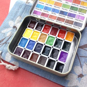 Puede incluir: Un juego de pintura de acuarela en una lata de metal, con una variedad de colores vibrantes. La lata est&aacute; abierta, revelando pozos de pintura individuales llenos de tonos como azul, amarillo, rojo y verde. Una bolsa de tela azul con un dise&ntilde;o floral est&aacute; al lado.