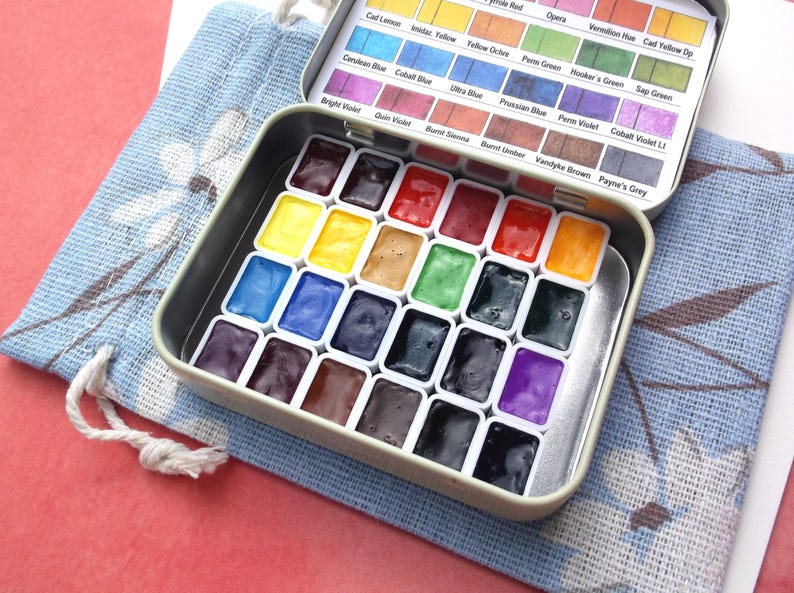 Puede incluir: Un juego de pintura de acuarela en una lata de metal, con una variedad de colores vibrantes. La lata est&aacute; abierta, revelando pozos de pintura individuales llenos de tonos como azul, amarillo, rojo y verde. Una bolsa de tela azul con un dise&ntilde;o floral est&aacute; al lado.