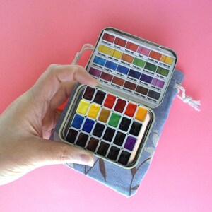 Puede incluir: Un peque&ntilde;o juego de pintura de acuarela de metal abierto con una variedad de colores, sostenido en una bolsa de tela azul. El juego incluye una carta de colores y una selecci&oacute;n de tonos vibrantes, ideal para artistas y aficionados.