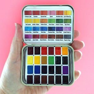 Puede incluir: Un peque&ntilde;o juego de pintura de acuarela abierto en una lata de metal, sostenido en una mano. La lata contiene 24 pastillas de pintura rectangulares en una variedad de colores, incluyendo rojo, amarillo, azul y verde. La tapa de la lata tiene los nombres de los colores.
