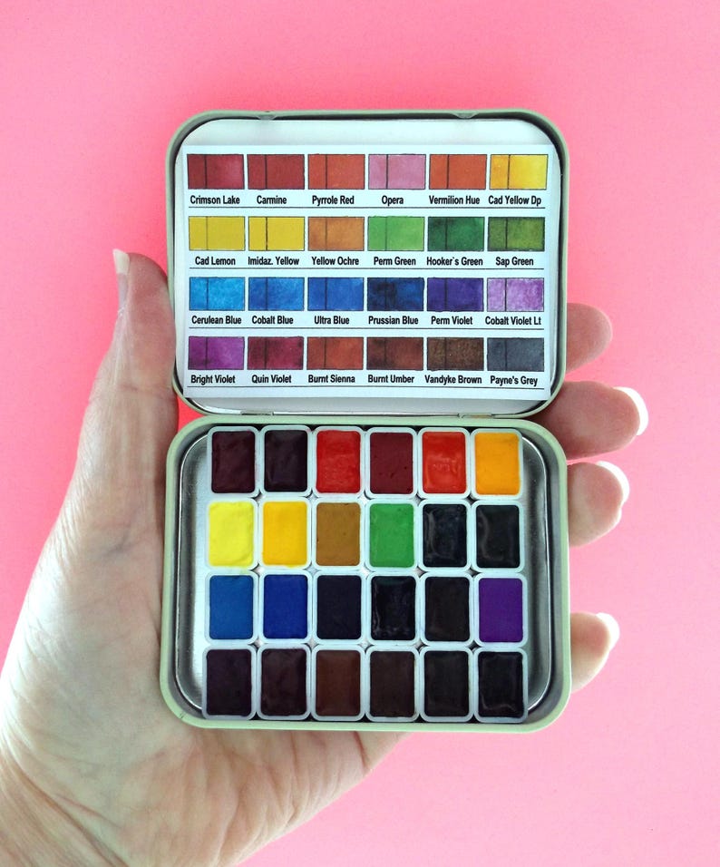 Puede incluir: Un peque&ntilde;o juego de pintura de acuarela abierto en una lata de metal, sostenido en una mano. La lata contiene 24 pastillas de pintura rectangulares en una variedad de colores, incluyendo rojo, amarillo, azul y verde. La tapa de la lata tiene los nombres de los colores.