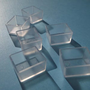 May include: Six clear plastic furniture leg protectors, each with a square shape and a slightly raised lip around the edge. The protectors are arranged in a triangular pattern on a blue surface.