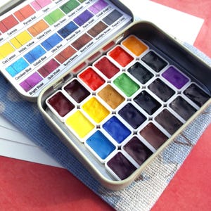 Puede incluir: Un juego de pintura de acuarela en una caja met&aacute;lica rectangular. La caja est&aacute; abierta, revelando filas de cuadrados de pintura coloridos y una tapa con nombres de colores. El juego descansa sobre un pa&ntilde;o azul y blanco.