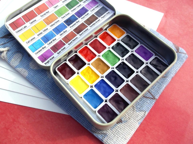 Puede incluir: Un juego de pintura de acuarela en una caja met&aacute;lica rectangular. La caja est&aacute; abierta, revelando filas de cuadrados de pintura coloridos y una tapa con nombres de colores. El juego descansa sobre un pa&ntilde;o azul y blanco.