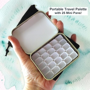 May include: Open portable travel watercolor palette with 25 mini pans. The rectangular tin case is held open, revealing the empty white pans. The text "Portable Travel Palette with 25 Mini Pans!" is displayed above the palette.