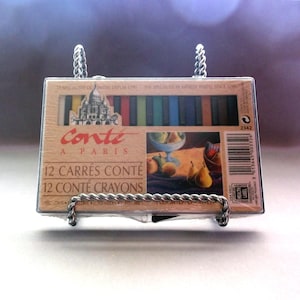 May include: A box of Conté à Paris 12 Carrés Conté crayons, displayed in a decorative metal holder. The box features a painting of fruit and a church, with text in French. The crayons are visible inside the box.