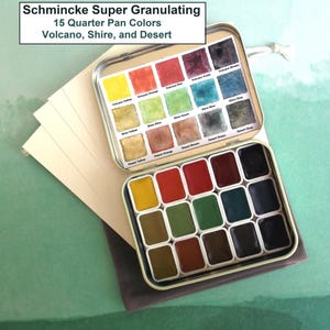 May include: A set of Schmincke Super Granulating watercolor paints in a metal tin. The tin contains 15 quarter pan colors, including Volcano, Shire, and Desert shades. The open tin is next to a stack of cream-colored paper.