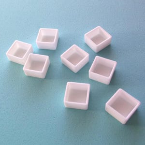 May include: Eight white square watercolor paint pans, arranged in a star shape on a light blue surface.