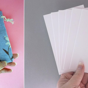 Puede incluir: Una bolsa azul con cord&oacute;n y un estampado floral, junto a una pila de tarjetas rectangulares blancas. La bolsa presenta flores blancas y tallos marrones. Las tarjetas se sostienen en una mano y la bolsa en otra. El fondo es rosa y gris.