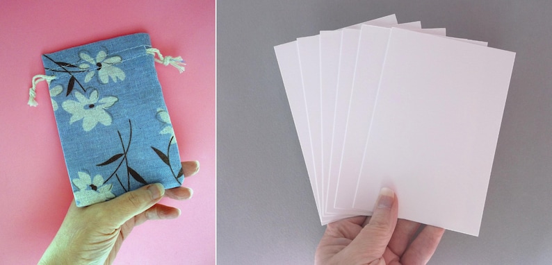 Puede incluir: Una bolsa azul con cord&oacute;n y un estampado floral, junto a una pila de tarjetas rectangulares blancas. La bolsa presenta flores blancas y tallos marrones. Las tarjetas se sostienen en una mano y la bolsa en otra. El fondo es rosa y gris.