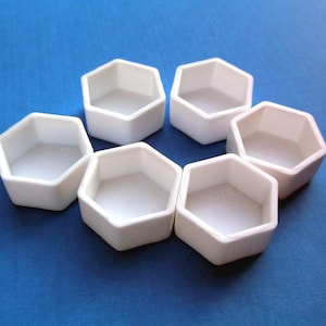 May include: Seven white, hexagonal ceramic containers are arranged in a circle on a blue surface. Each container has a flat bottom and angled sides. The containers are empty and appear to be new.