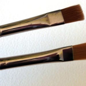 May include: Two flat makeup brushes with silver handles and brown bristles. The brushes are different sizes.