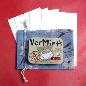 Puede incluir: Una bolsa azul floral con cord&oacute;n contiene una lata rectangular de pastillas de chai VerMints, con una ilustraci&oacute;n de taza de t&eacute;. La lata est&aacute; etiquetada como "VerMints All Natural Chai Pastilles" y "Peso neto 40g". Varias tarjetas blancas son visibles detr&aacute;s de la bolsa.