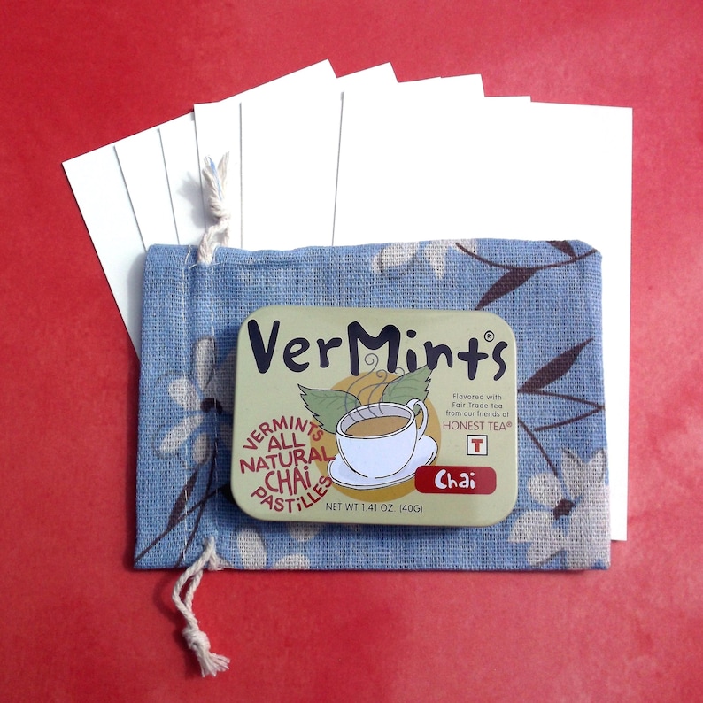 Puede incluir: Una bolsa azul floral con cord&oacute;n contiene una lata rectangular de pastillas de chai VerMints, con una ilustraci&oacute;n de taza de t&eacute;. La lata est&aacute; etiquetada como "VerMints All Natural Chai Pastilles" y "Peso neto 40g". Varias tarjetas blancas son visibles detr&aacute;s de la bolsa.