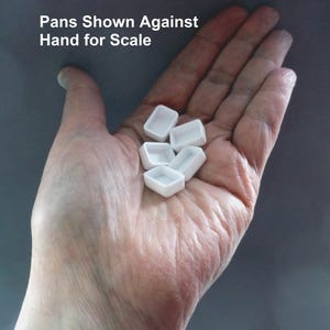 Puede incluir: Una mano que sostiene varios godets de pintura de acuarela peque&ntilde;os, blancos y rectangulares. Los godets est&aacute;n vac&iacute;os y parecen ser de cer&aacute;mica o porcelana. El texto de la imagen dice "Pans Shown Against Hand for Scale". El fondo es gris liso.
