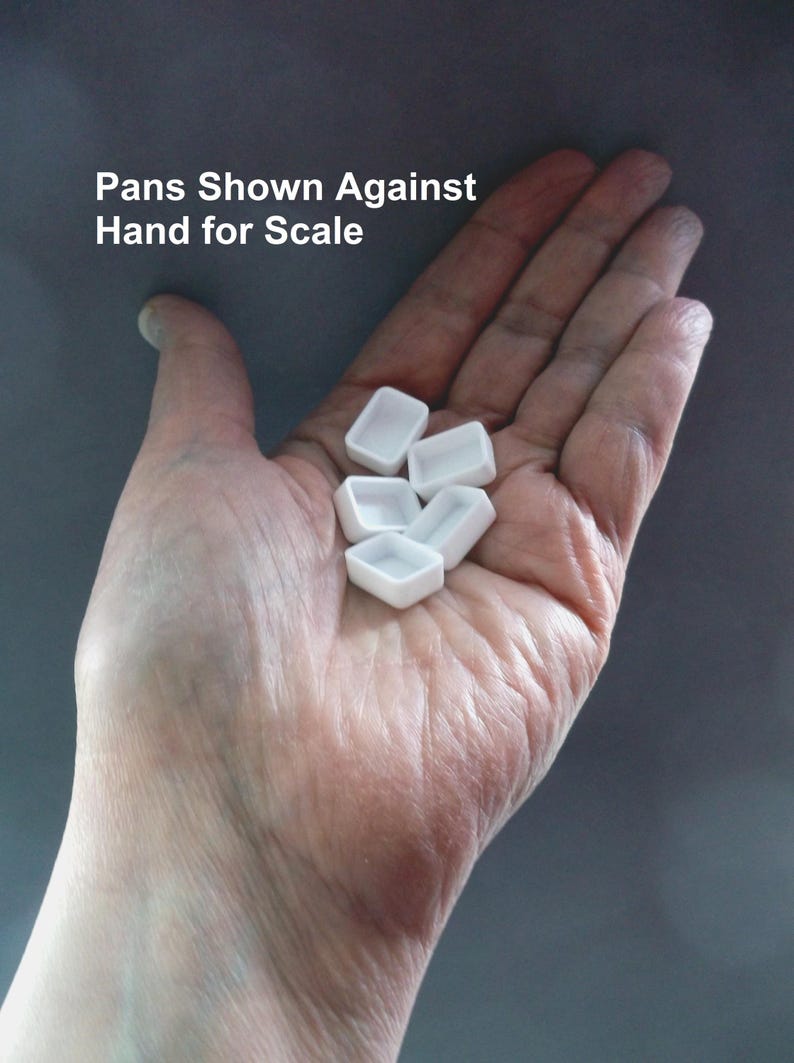 Puede incluir: Una mano que sostiene varios godets de pintura de acuarela peque&ntilde;os, blancos y rectangulares. Los godets est&aacute;n vac&iacute;os y parecen ser de cer&aacute;mica o porcelana. El texto de la imagen dice "Pans Shown Against Hand for Scale". El fondo es gris liso.