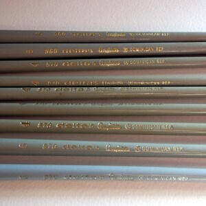 May include: A close-up of a stack of gray pencils with gold lettering. The pencils are labeled with the text "delta's Graphite DOMINICAN REP" and numbers ranging from 820 to 970.