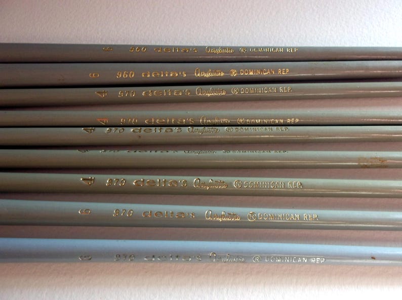 May include: A close-up of a stack of gray pencils with gold lettering. The pencils are labeled with the text "delta's Graphite DOMINICAN REP" and numbers ranging from 820 to 970.