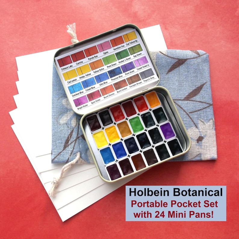 Puede incluir: Un juego de acuarelas de bolsillo port&aacute;til Holbein Botanical con 24 mini godets. La lata abierta presenta una variedad de colores vibrantes. El juego se muestra sobre una tela azul con estampado floral y papel blanco.