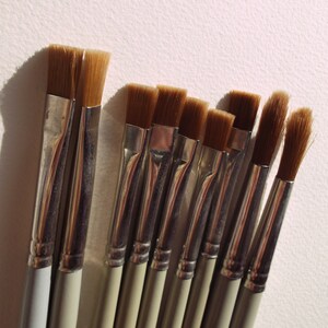 May include: A set of nine paint brushes with brown bristles and silver handles. The brushes are arranged in a row, with the largest brush on the left and the smallest brush on the right.