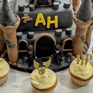 May include: A two-tiered cake shaped like a castle with gray turrets and brown walls. The cake is decorated with the letters "AH" in yellow. Cupcakes with yellow frosting and gold toppers surround the cake.