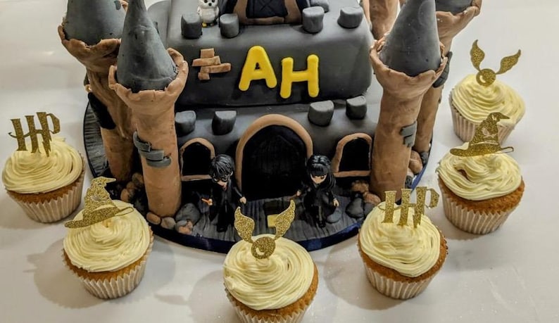 May include: A two-tiered cake shaped like a castle with gray turrets and brown walls. The cake is decorated with the letters "AH" in yellow. Cupcakes with yellow frosting and gold toppers surround the cake.