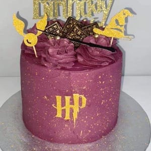 May include: A round, burgundy cake with gold accents and decorations. The cake is topped with burgundy frosting, gold glitter accents, and Harry Potter-themed toppers, including a golden snitch, sorting hat, and the words "Happy Birthday".