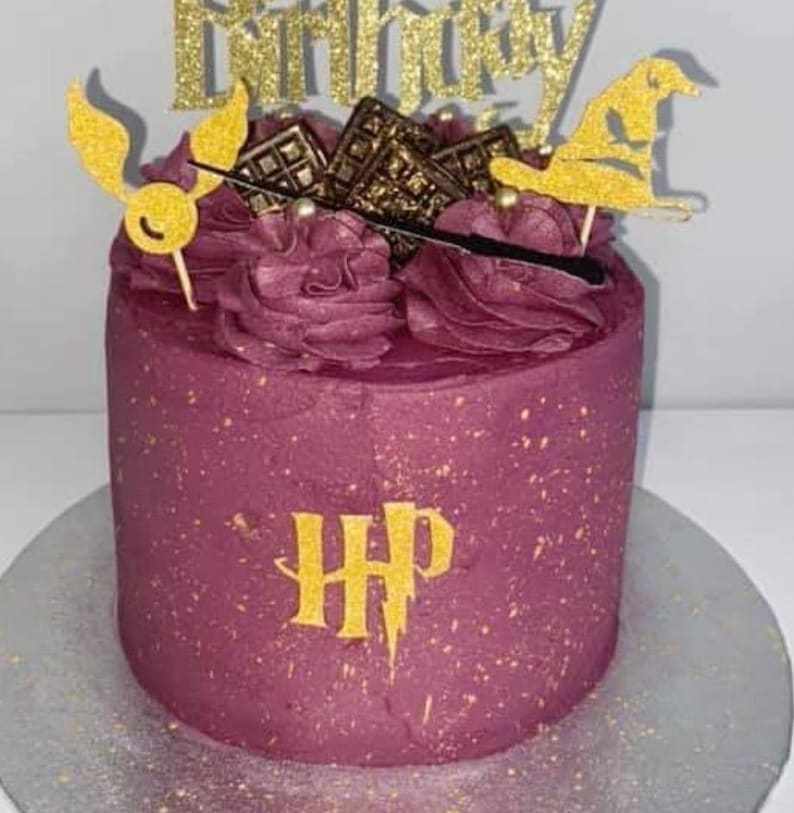 May include: A round, burgundy cake with gold accents and decorations. The cake is topped with burgundy frosting, gold glitter accents, and Harry Potter-themed toppers, including a golden snitch, sorting hat, and the words "Happy Birthday".