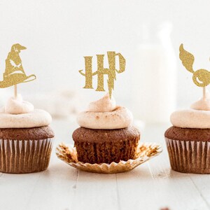 May include: Three chocolate cupcakes with light brown frosting and gold glitter toppers. The toppers include a wizard hat, the letters "HP", and a winged golden ball. The cupcakes sit on a white surface.