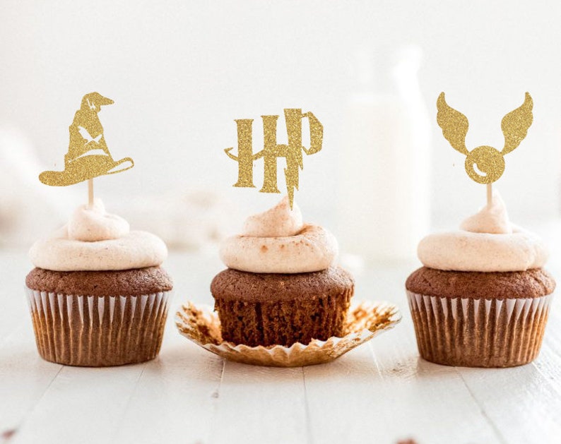 May include: Three chocolate cupcakes with light brown frosting and gold glitter toppers. The toppers include a wizard hat, the letters "HP", and a winged golden ball. The cupcakes sit on a white surface.
