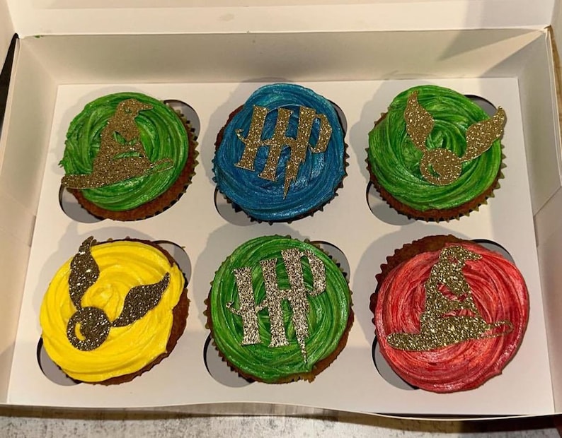 May include: Six cupcakes in a white box, each with colorful frosting and gold glitter decorations. The cupcakes have green, blue, yellow, and red frosting. The decorations include a wizard hat, lightning bolt, and golden snitch.