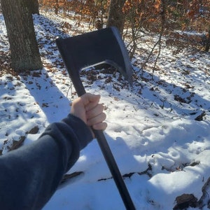 May include: A black axe with a wooden handle, held by a person in a snowy forest setting.