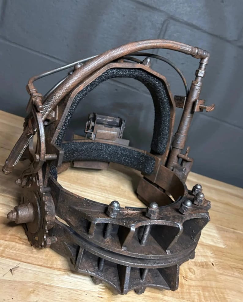 K&ouml;nnte beinhalten: Eine rostige Metall-Kopf-Falle mit einem gepolsterten Innenraum und einer Reihe von Zahnr&auml;dern und Schrauben. Die Falle ist so konzipiert, dass sie um den Kopf einer Person passt und eine scharfe, gezackte Kante hat.