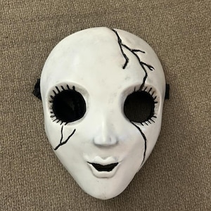 May include: White porcelain mask with black painted eyes, eyelashes, and a cracked design. The mask has a wide, open mouth.