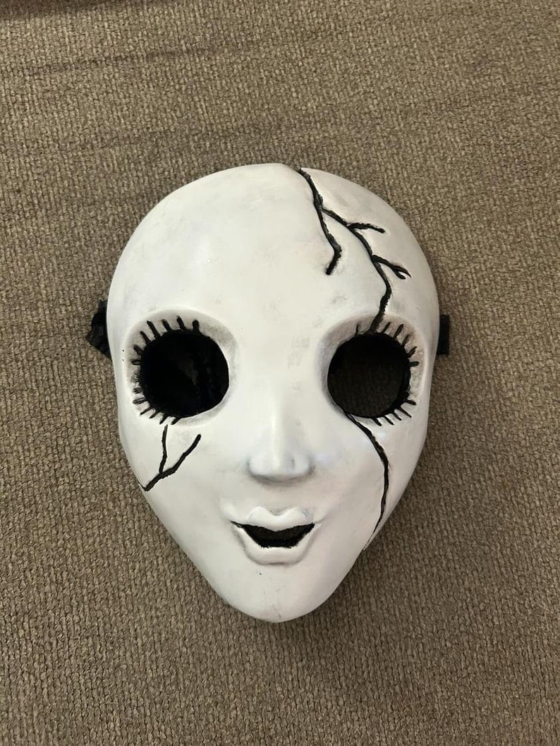 May include: White porcelain mask with black painted eyes, eyelashes, and a cracked design. The mask has a wide, open mouth.