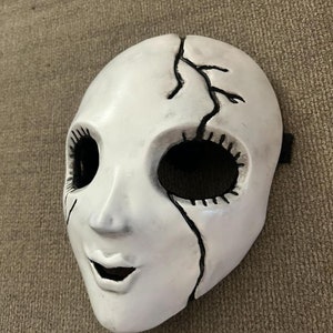 May include: White porcelain mask with a cracked design and black painted details. The mask has a wide-eyed expression with a stitched mouth.