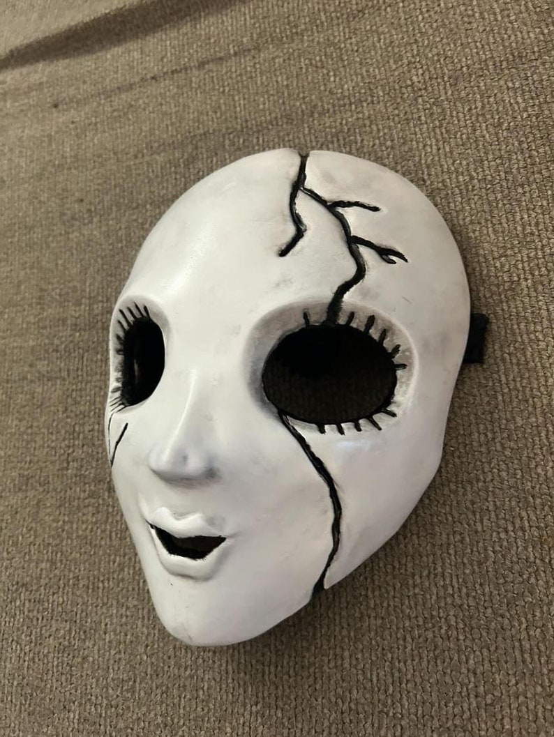 May include: White porcelain mask with a cracked design and black painted details. The mask has a wide-eyed expression with a stitched mouth.
