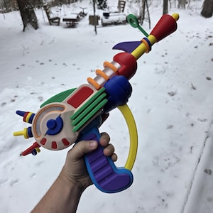 May include: A brightly coloured toy ray gun, featuring red, blue, green, yellow, and purple elements. The ray gun has a circular front with small protrusions and a handle with purple ridges. The background shows a snowy outdoor scene.