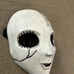 May include: White porcelain doll mask with black painted eyes, mouth, and stitching details. The mask has a cracked and distressed look.