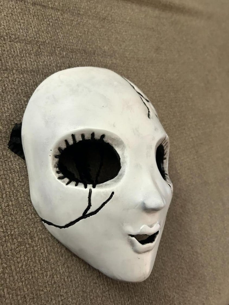 May include: White porcelain doll mask with black painted eyes, mouth, and stitching details. The mask has a cracked and distressed look.