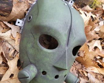 Könnte beinhalten: Eine grüne Hockeymaske mit einer abgenutzten Lackierung und Metallverzierungen. Die Maske hat ein großes, rundes Augenloch und ein kleineres, rechteckiges Mundloch. Die Maske besteht aus hartem Kunststoff und hat eine strukturierte Oberfläche.